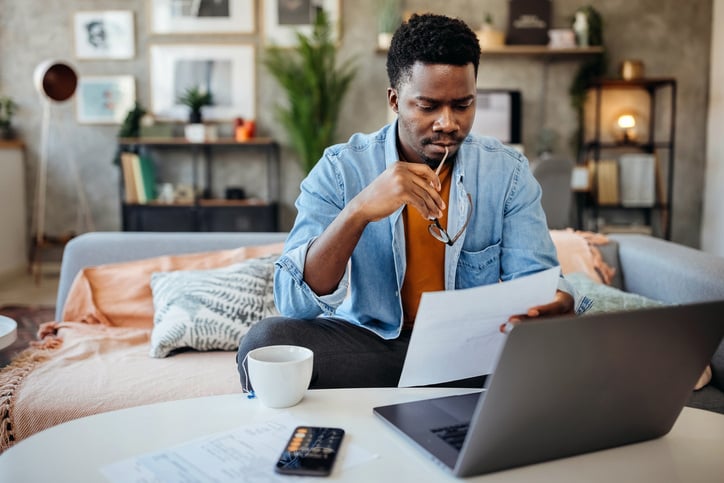 a-man-sits-at-a-table-with-the-end-of-his-glasses-in-his-mouth-while-looking-at-a-piece-of-paper-with-his-laptop-and-calculator-out