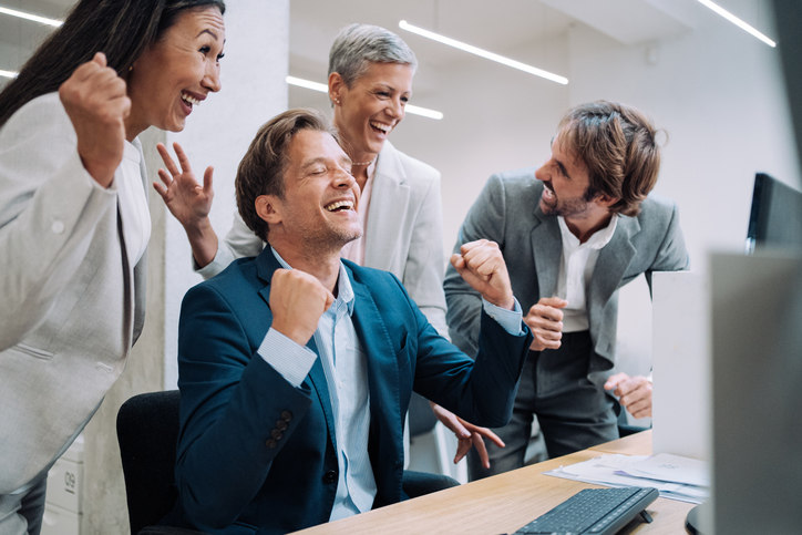 a-group-of-people-stand-around-a-man-at-a-desk-and-celebrate