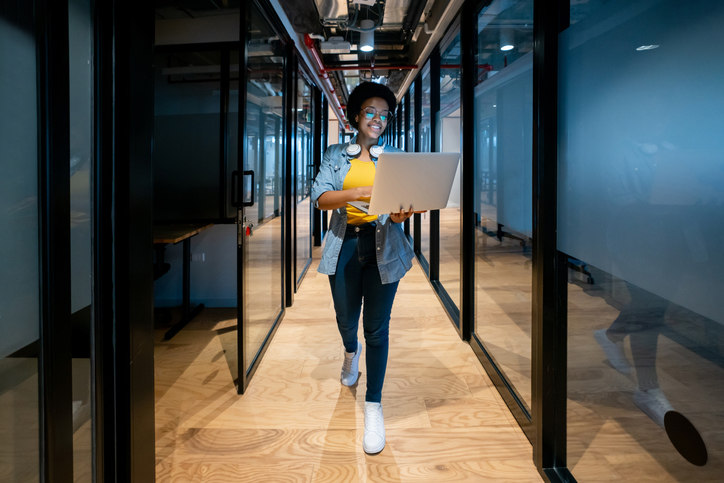 woman-walking-down-a-hallway-while-working-on-her-laptop