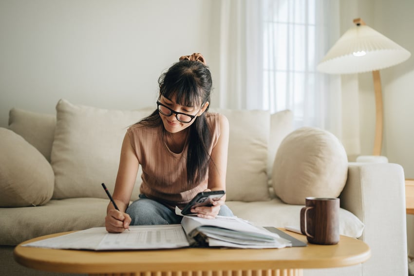 a-girl-sitting-at-a-couch-working-on-a-piece-of-paper-while-looking-at-her-phone