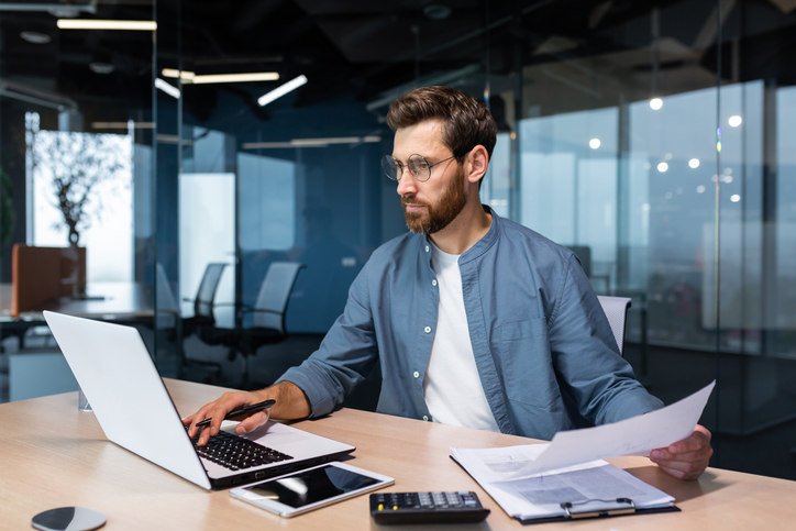 a-man-sits-at-a-desk-and-types-information-from-a-piece-of-paper-into-a-computer