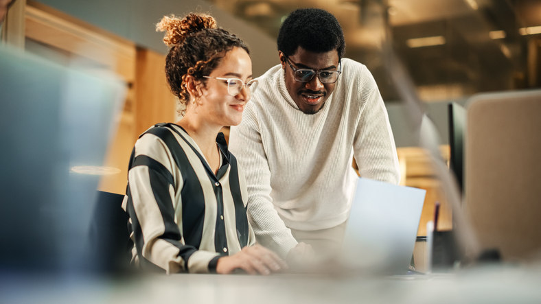 a-woman-and-a-man-look-at-a-computer-with-the-woman-sitting-and-the-man-standing