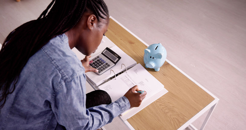 a-woman-sits-at-a-desk-at-writes-down-information-on-a-piece-of-paper-while-typing-numbers-into-a-calculator