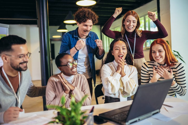 a-group-of-people-stand-around-a-laptop-cheering