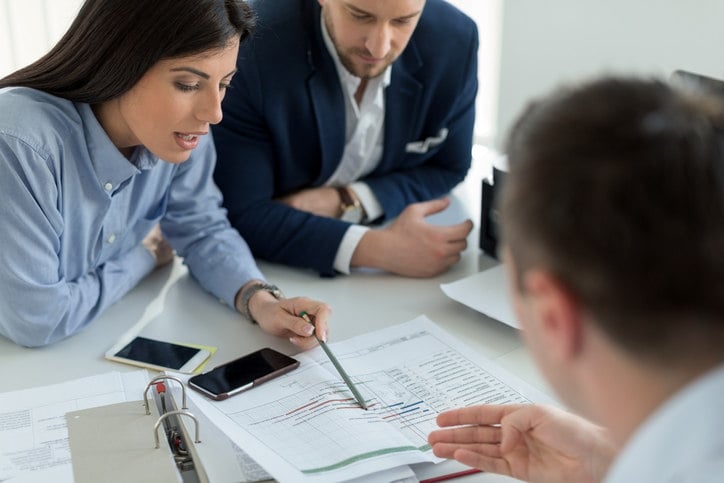 three-people-gathered-around-a-table-looking-at-a-piece-of-paper-with-data